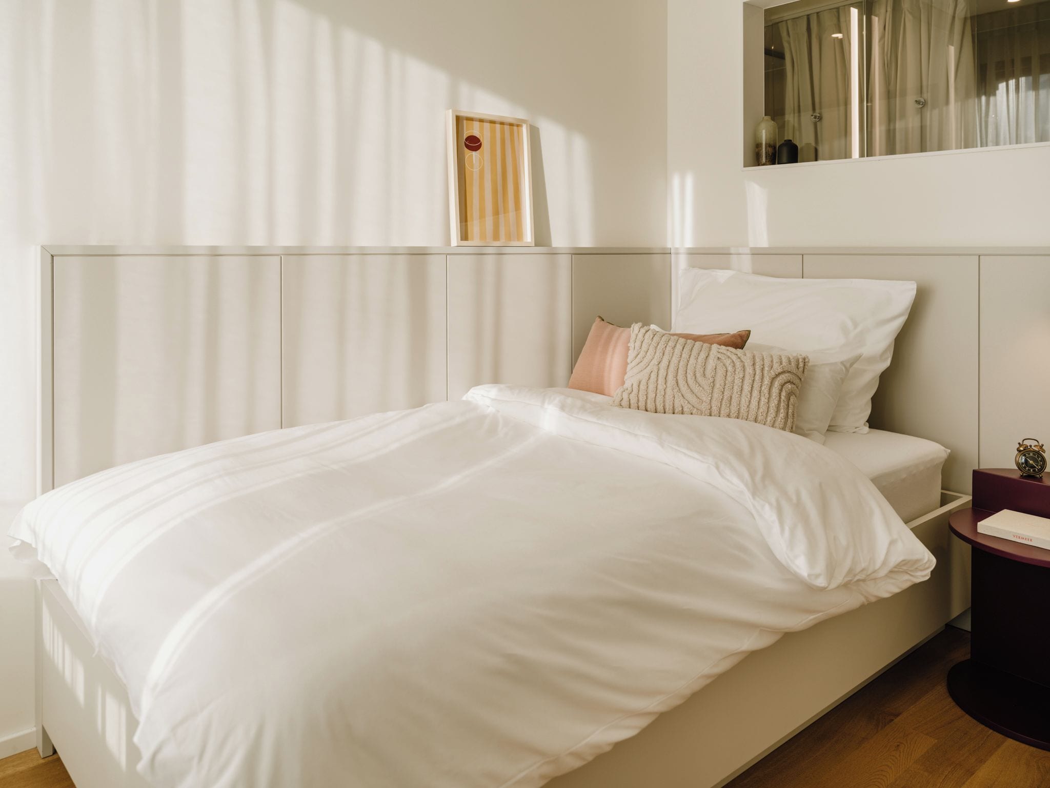 Una cama bien hecha con sábanas blancas, una mesita de noche con un libro y un despertador, y una ventana con cortinas translúcidas.