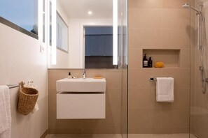 Ein modernes Badezimmer mit einer gläsernen Dusche, einem weißen Waschbecken mit Spiegel darüber und einem Handtuchhalter mit einem gewebten Korb.