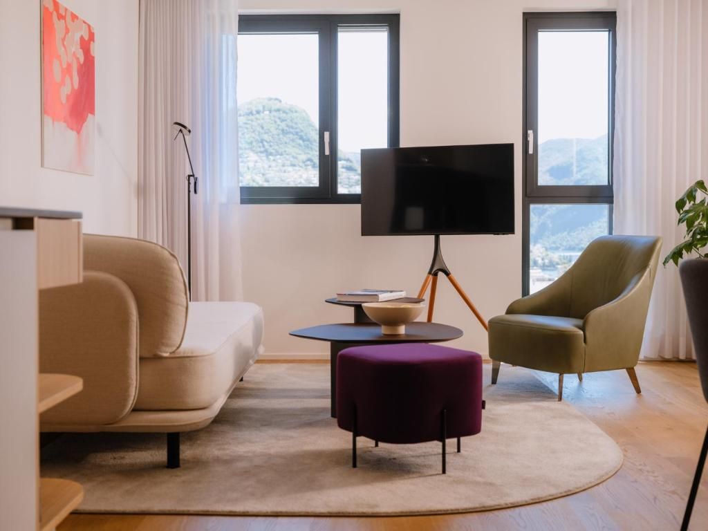 Uma sala de estar moderna com sofá, banqueta e poltrona, uma TV de tela plana e vista para o exterior por grandes janelas.