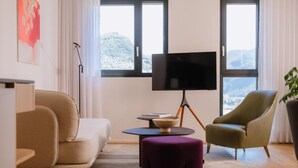 A modern living room with a sofa, ottoman, and armchair, a flat-screen TV, and a view of the outdoors through large windows. - THE FLAG Lugano Self Check-in (Massagno)