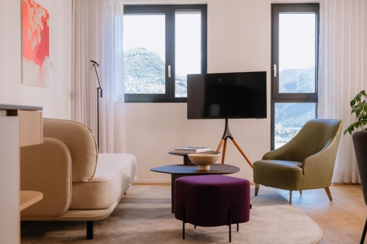 A modern living room with a sofa, ottoman, and armchair, a flat-screen TV, and a view of the outdoors through large windows.