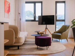 Ein modernes Wohnzimmer mit einer Couch, Hocker und Sesseln, einem Flachbildfernseher und Blick ins Freie durch große Fenster.