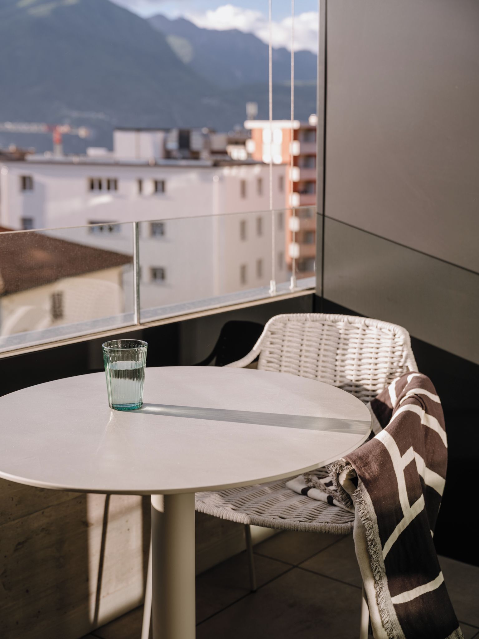 Ein Balkon mit rundem Tisch und einem Stuhl, einem Glas Wasser und Blick auf Gebäude und Berge.