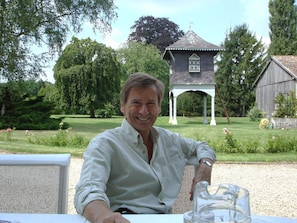 A man seated at an outdoor table with a clear view of a gazebo and a barn in the background.