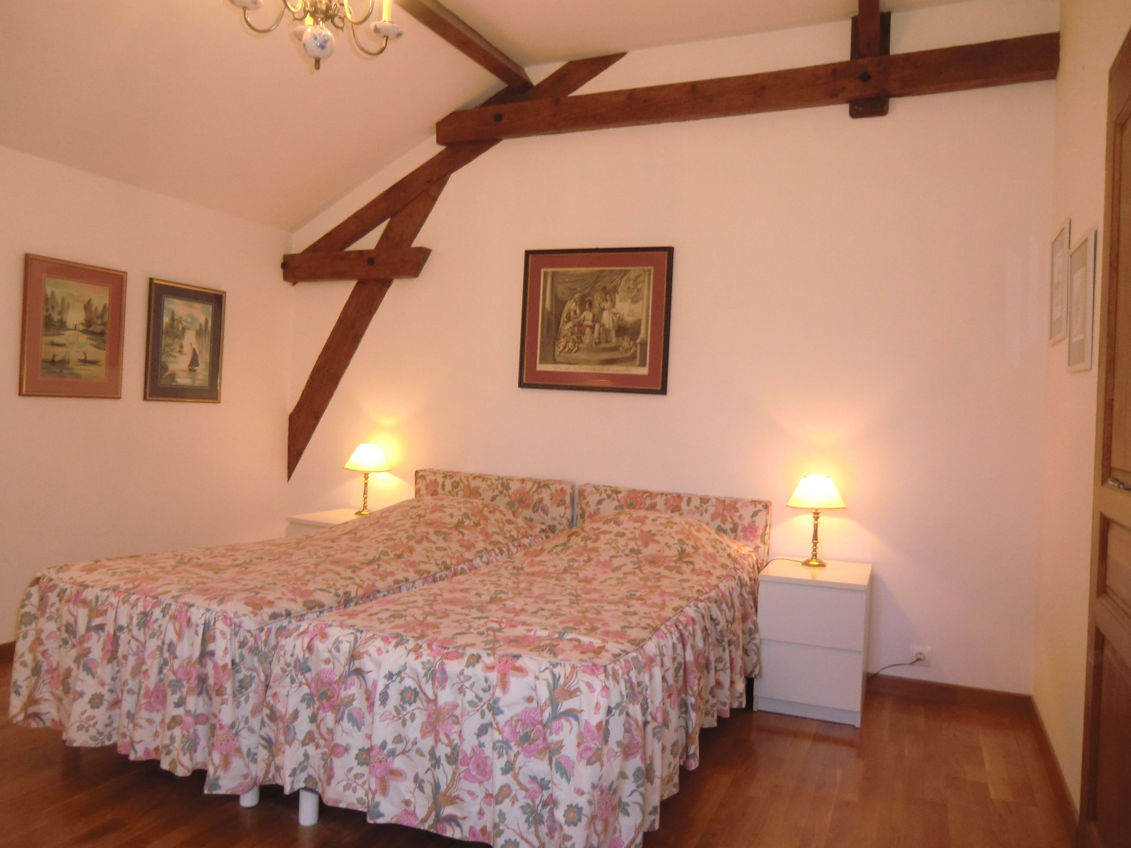 Ein Schlafzimmer mit Holzbalkendecke, einer blumigen Bettdecke, zwei Nachttischlampen und zwei gerahmten Bildern an der Wand.