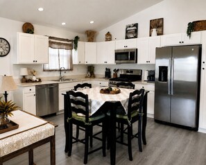 Une cuisine moderne avec des armoires blanches, des appareils en acier inoxydable, une table à manger avec des chaises et une horloge au mur.
