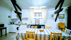 Uma sala de jantar com mesa posta para quatro pessoas, cadeiras de madeira, cozinha ao fundo e janela com cortinas.
