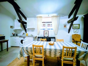 Een eetgedeelte met een tafel voor vier personen, houten stoelen, een keuken op de achtergrond en een raam met gordijnen.