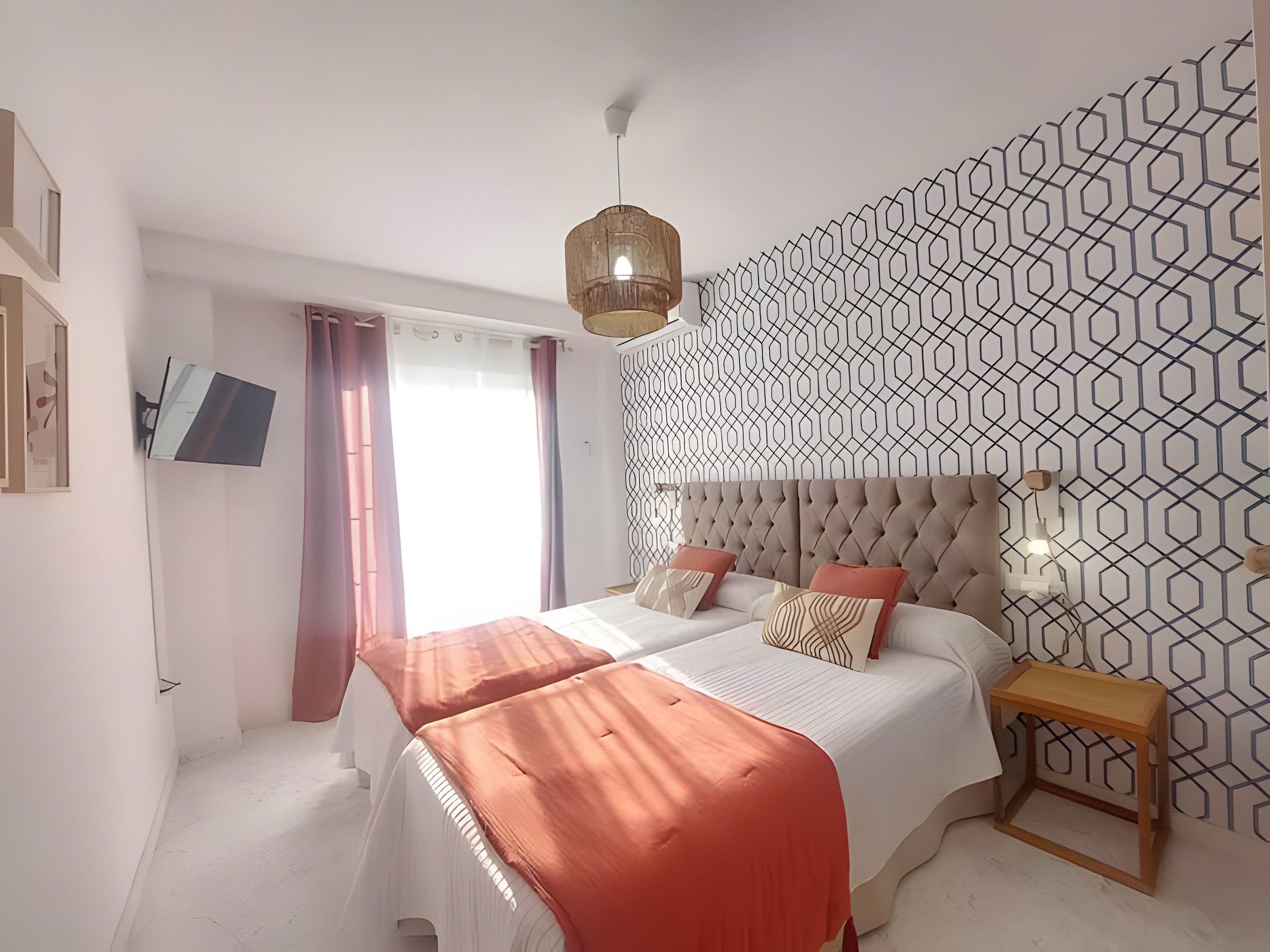 Un dormitorio con una cama, una mesita de noche, un televisor montado en la pared y un papel tapiz estampado.