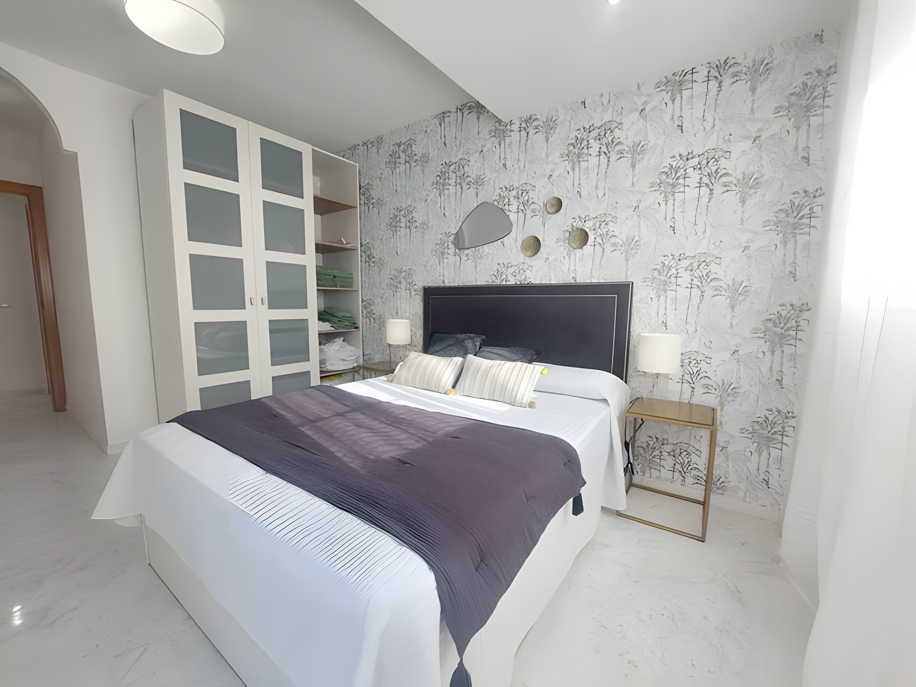 Un dormitorio con una cama, un armario de puertas de cristal, una mesita de noche y una pared con un diseño de árbol.