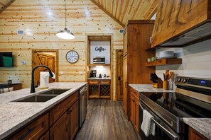 A kitchen with wooden walls, a clock, a coffee sign, and modern appliances. - 1BR Secluded Hot Tub Retreat (Broken Bow)