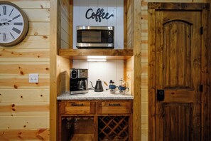 A kitchen with wooden walls, a clock, a coffee sign, a microwave, a coffee maker, and a wine cooler. - 1BR Secluded Hot Tub Retreat (Broken Bow)