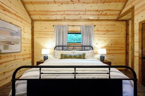 A wooden cabin bedroom with a metal bed frame, two bedside lamps, and a window with curtains. - 1BR Secluded Hot Tub Retreat (Broken Bow)