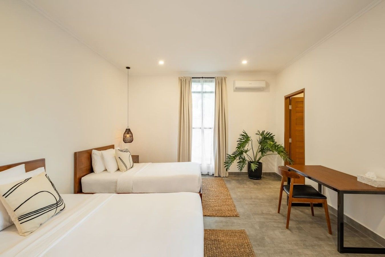 Une chambre d’hôtel avec deux lits, un bureau, une chaise, une plante en pot et une fenêtre avec des rideaux.