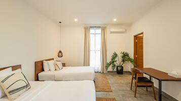 Une chambre d’hôtel avec deux lits, un bureau, une chaise, une plante en pot et une fenêtre avec des rideaux.