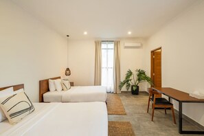 Une chambre d’hôtel avec deux lits, un bureau, une chaise, une plante en pot et une fenêtre avec des rideaux.