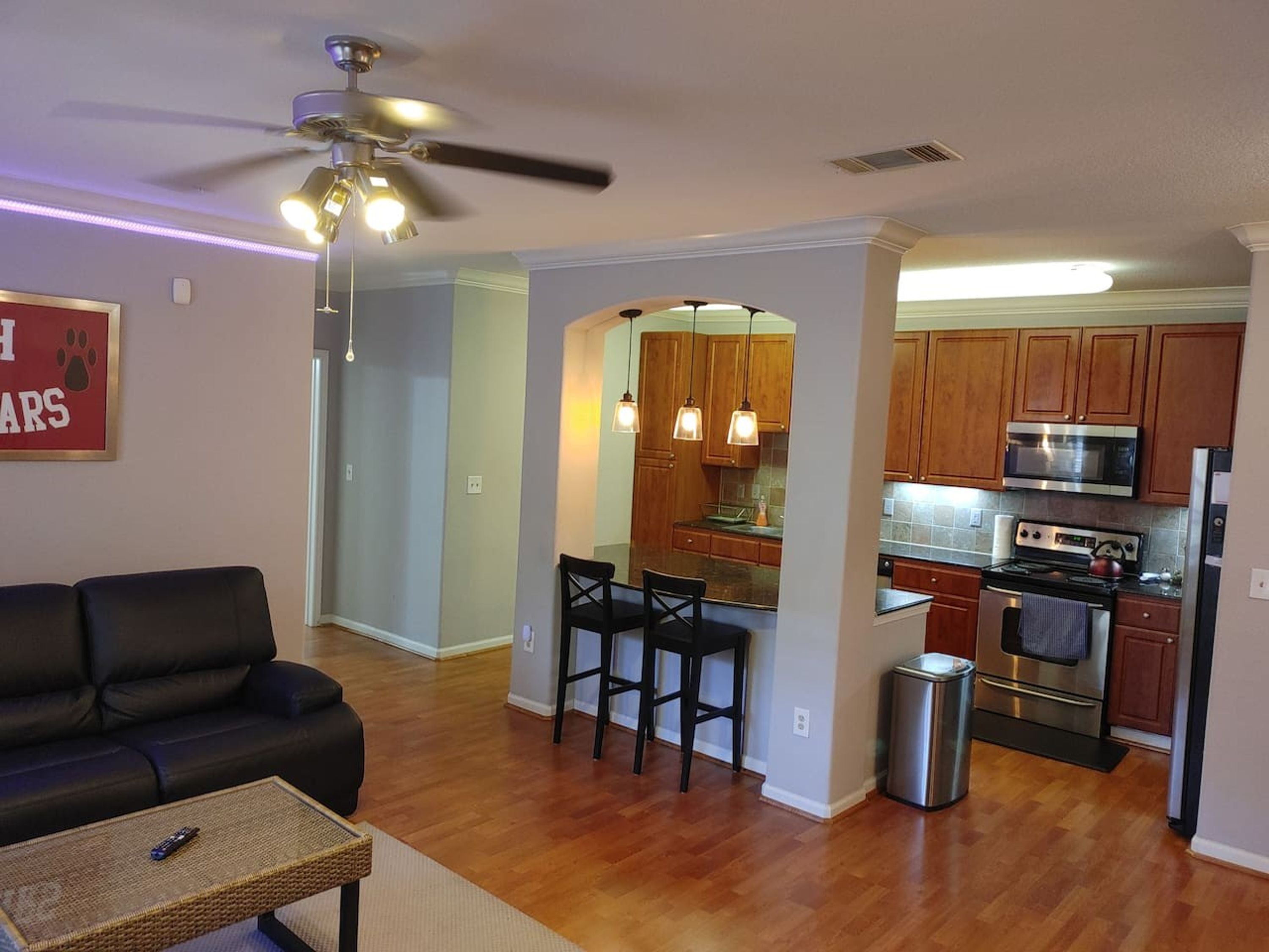 A living room with a black sofa, a coffee table, and a ceiling fan. There is a kitchen area with wooden cabinets, a microwave, and a stove.