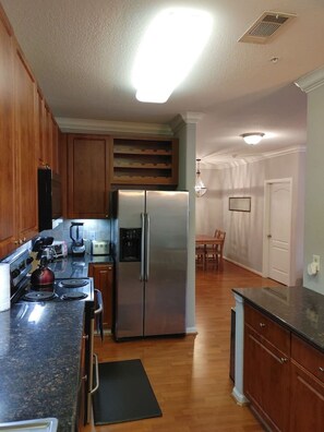 A kitchen with wooden cabinets, a stainless steel refrigerator, a granite countertop, and a dining area with a table and chairs. - Med Center Condo With Pool Gym and Balcony (Houston)