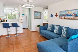 Ein modernes Wohnzimmer mit einem blauen Sofa, zwei Barhocker, einem Essbereich mit einem weißen Tisch und einer Küche im Hintergrund.