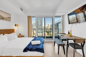 Een moderne hotelkamer met een groot bed, een eettafel, een bankje en stadszicht door raam-tot-vloer ramen.