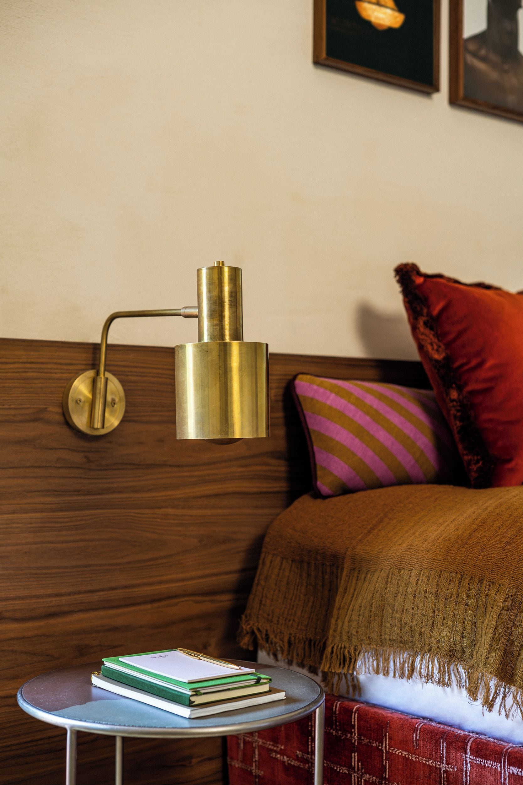 Un lit avec une tête de lit en bois, une lampe de chevet, une petite table ronde avec des livres et un coussin coloré.