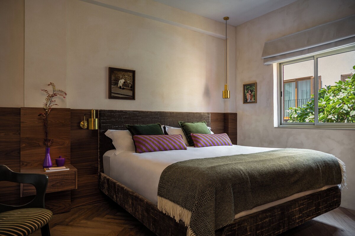 A bedroom with a large bed, a wooden headboard, a nightstand, a chair, and a window with a view of greenery.
