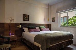 A bedroom with a large bed, a wooden headboard, a nightstand, a chair, and a window with a view of greenery. - The House of Delray (Tel Aviv)
