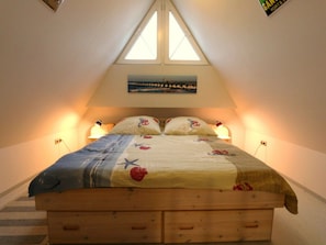 Een slaapkamer met een schuine kap, een bed met een bloemenprint deken, twee bedlampjes en een schilderij boven het bed.