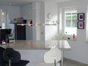 A modern kitchen with a central island, a dining area with chairs, a clock on the wall, and a window with a view. - Rosenberg Settlement 21 (Zingst)