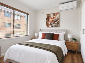 A bedroom with a bed, nightstands, a wall-mounted air conditioner, and a window with blinds. - Chic St Kilda retreat with secure parking (St Kilda)