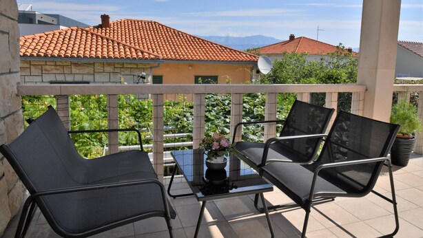 A balcony with two black chairs and a table, a potted plant, and a view of a red-tiled roof building. - A2 Apartment with terrace, 1 min walking to the beach (Supetar)
