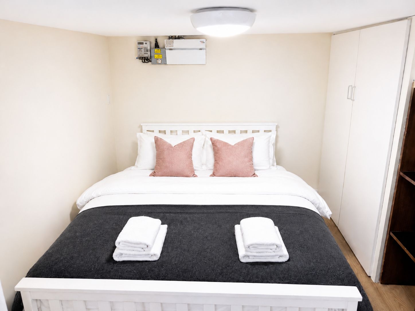 A neatly made bed with two pillows, a bedside table with a folded white towel, and a wall-mounted heater.