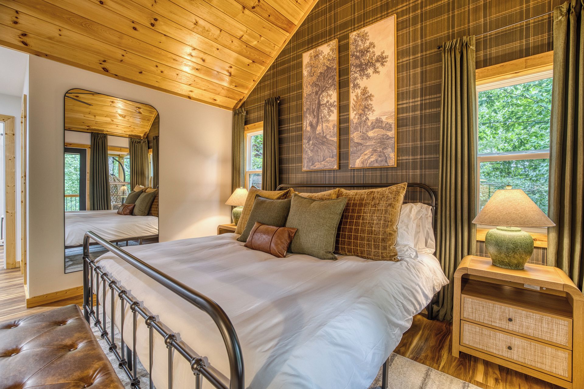 A bedroom with a wooden ceiling, a large bed with multiple pillows, a nightstand with a lamp, and a window with curtains.