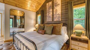 A bedroom with a wooden ceiling, a large bed with multiple pillows, a nightstand with a lamp, and a window with curtains.