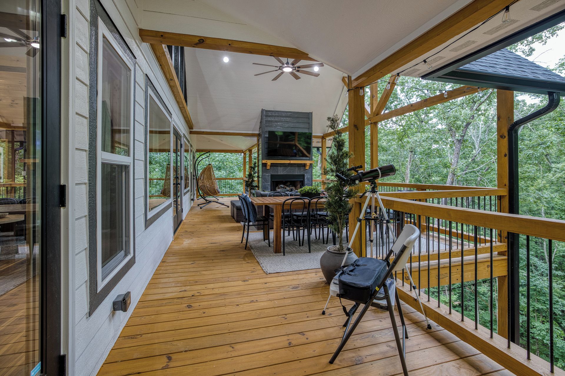 A covered deck with a wooden floor, a dining area with chairs and a table, a fireplace, and a telescope.