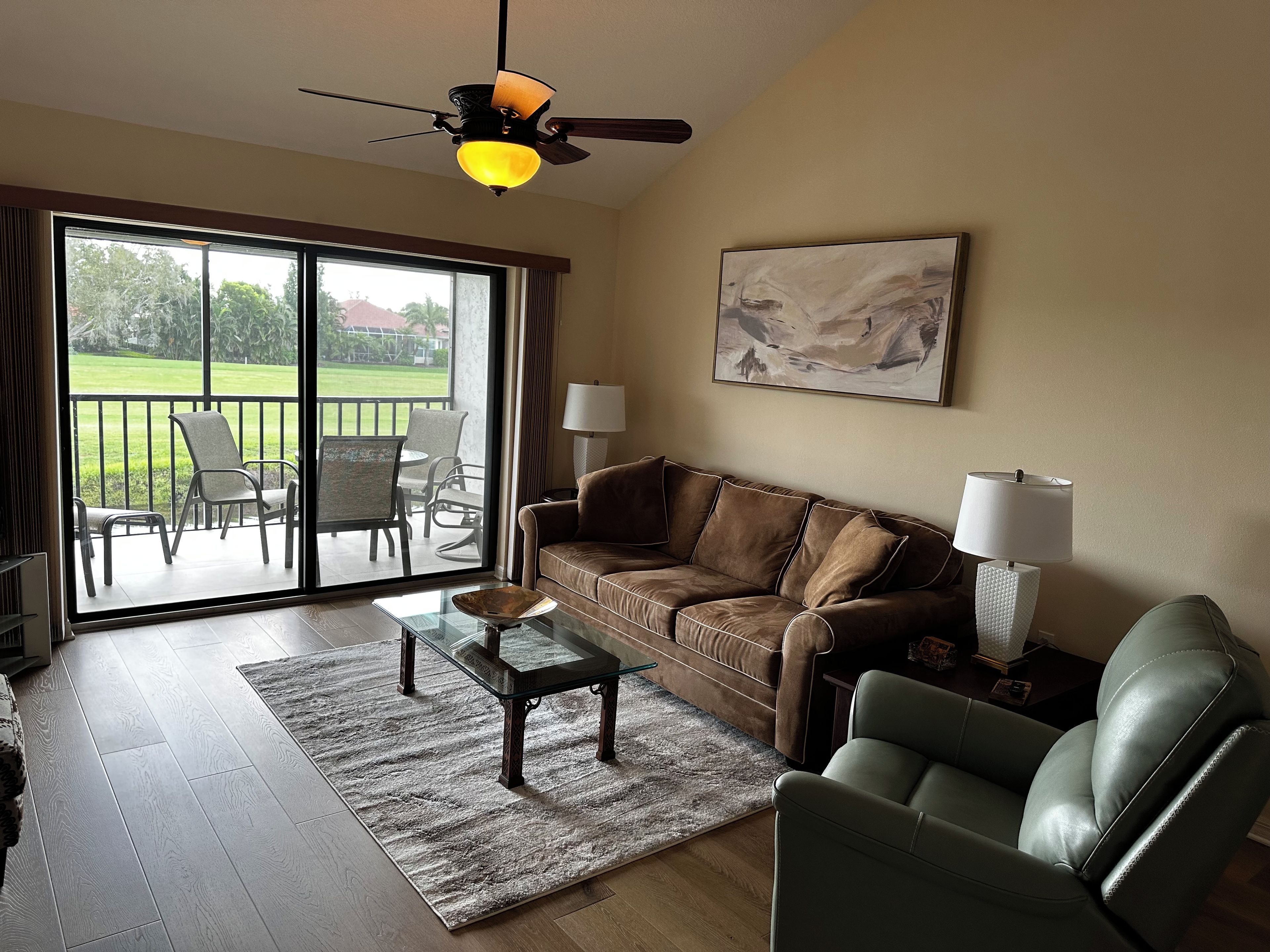 A living room with a brown sofa, a glass coffee table, a green armchair, a lamp, and a painting on the wall.