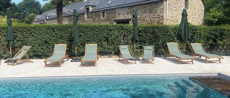 A poolside area with lounge chairs and umbrellas, a stone building in the background, and a clear blue pool.