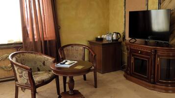 Habitación de hotel con una silla de madera, una mesita redonda con un libro, un mueble con televisor de pantalla plana y una ventana con cortinas.