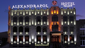 Ein Gebäude mit den Schildern 'ALEXANDRAPOL' und 'HOTEL PALACE', das einen zentralen Eingang mit einem dekorativen Bogen und mehrere beleuchtete Fenster zeigt.