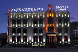 Een gebouw met de aanduiding 'ALEXANDRAPOL' en 'HOTEL PALACE', met een centrale ingang met een decoratieve boog en meerdere verlichte ramen.