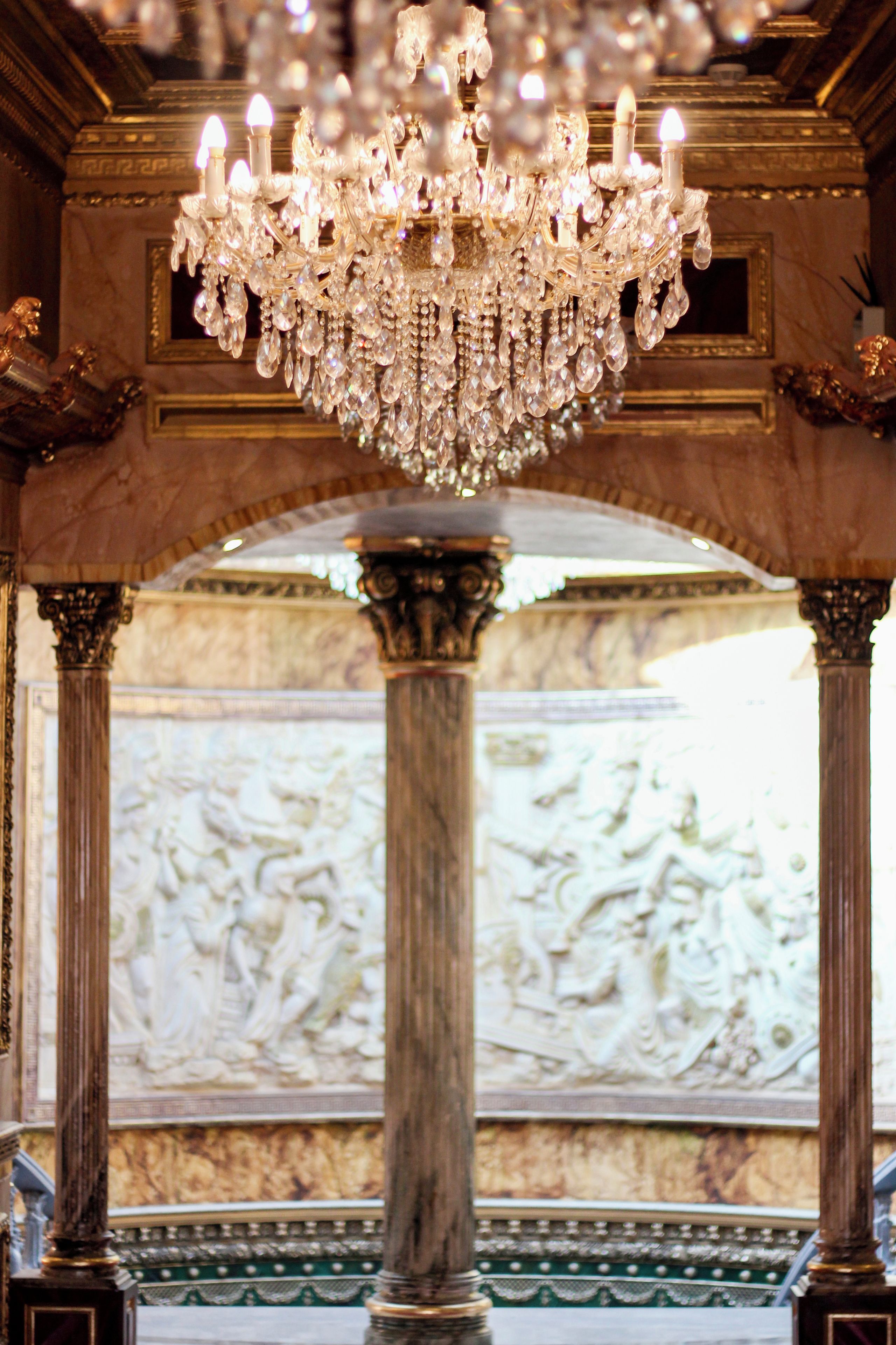 A grand chandelier with multiple lights, a marble column, and a classical architectural backdrop.