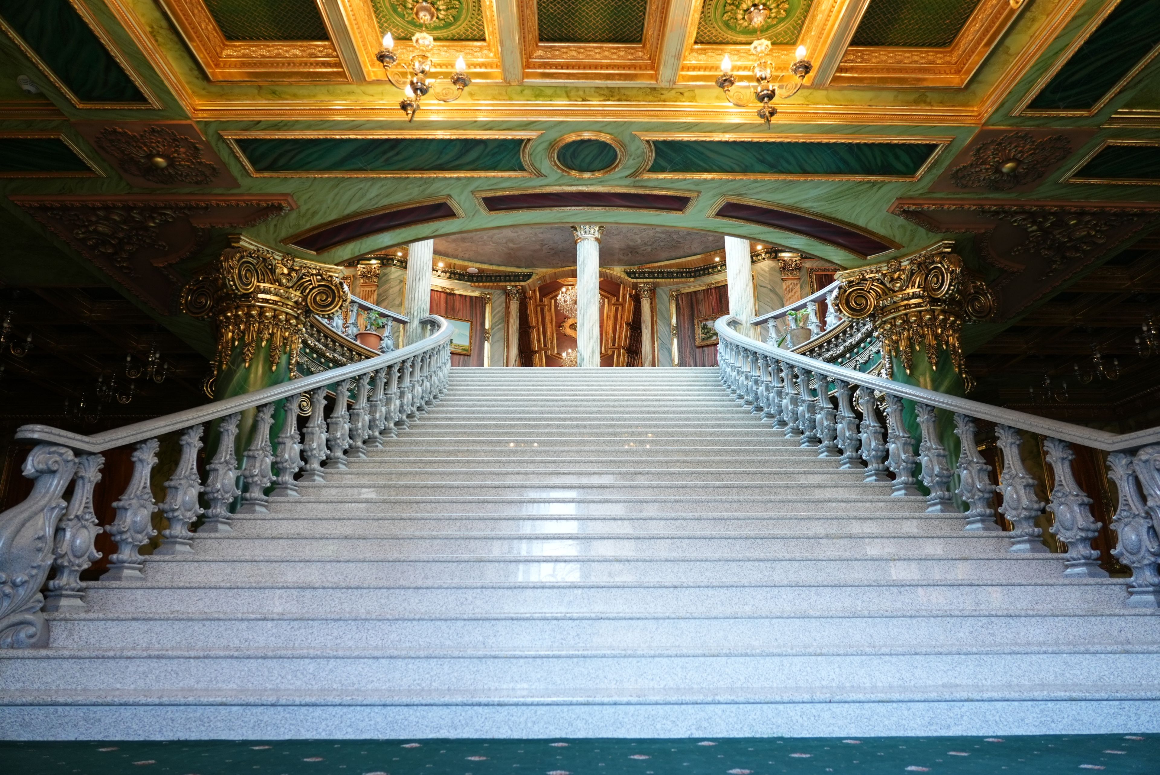 A grand staircase with ornate railings and a marble floor leading to a richly decorated room with a chandelier.