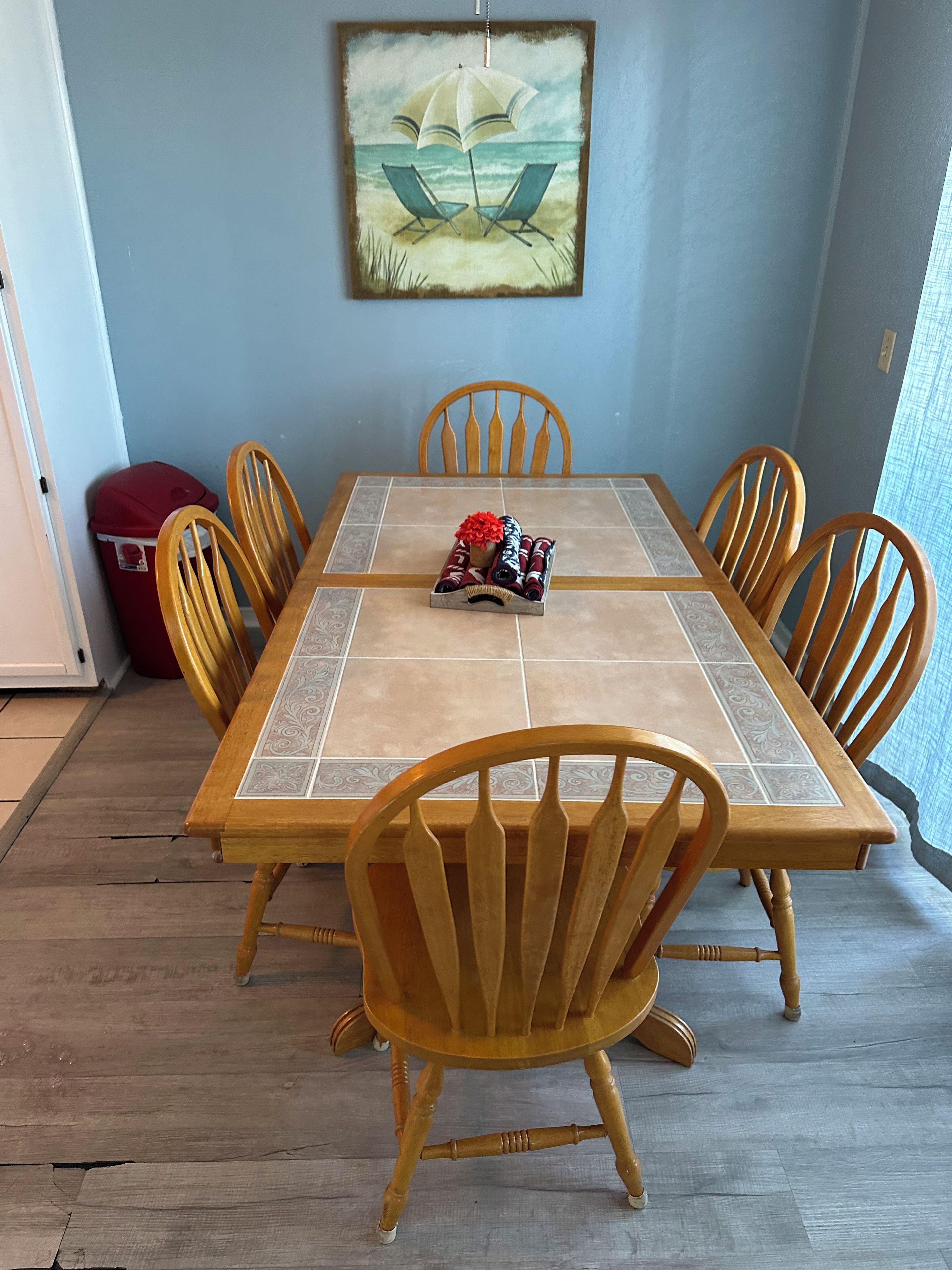 A dining area with a table set for six, surrounded by wooden chairs. A painting of a beach scene hangs on the wall above the table.