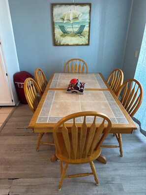 Ein Essbereich mit einem für sechs Personen gedeckten Tisch, umgeben von Holzstühlen. Über dem Tisch hängt ein Gemälde mit einer Strandansicht.