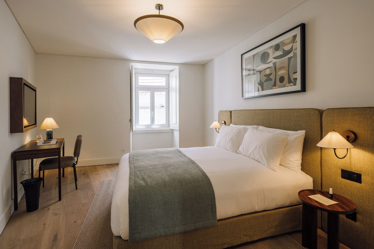 A hotel room with a bed, bedside tables, a desk, a chair, a window, and a painting on the wall.