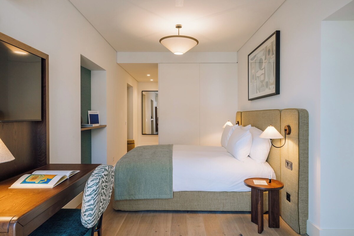 A hotel room with a bed, bedside lamps, a desk with a chair, a mirror, and a framed picture on the wall.