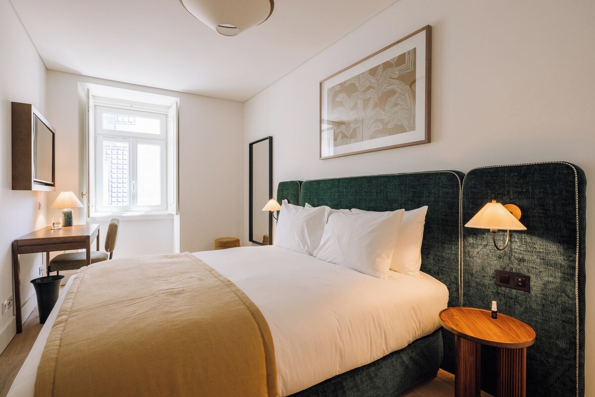 A hotel room with a bed, bedside tables, a desk, a chair, and a window.