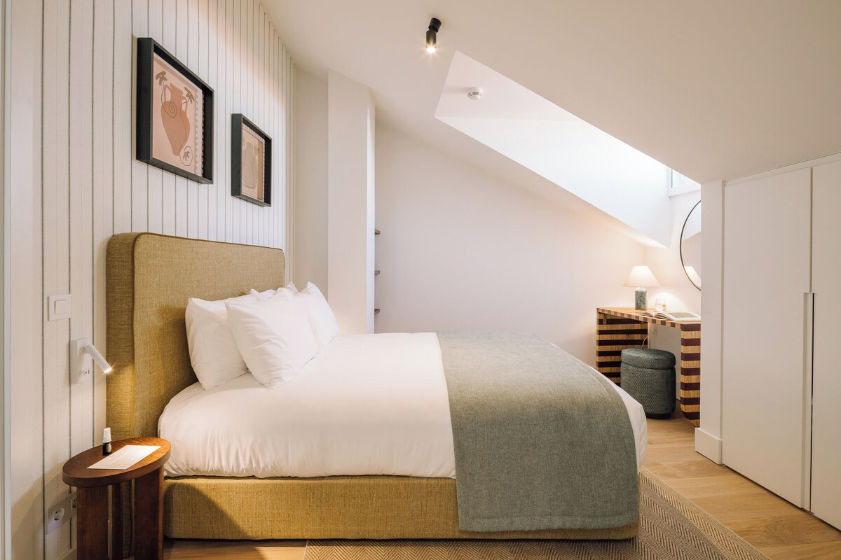 A bedroom with a bed, bedside table, lamp, and a view of the sky through a skylight.