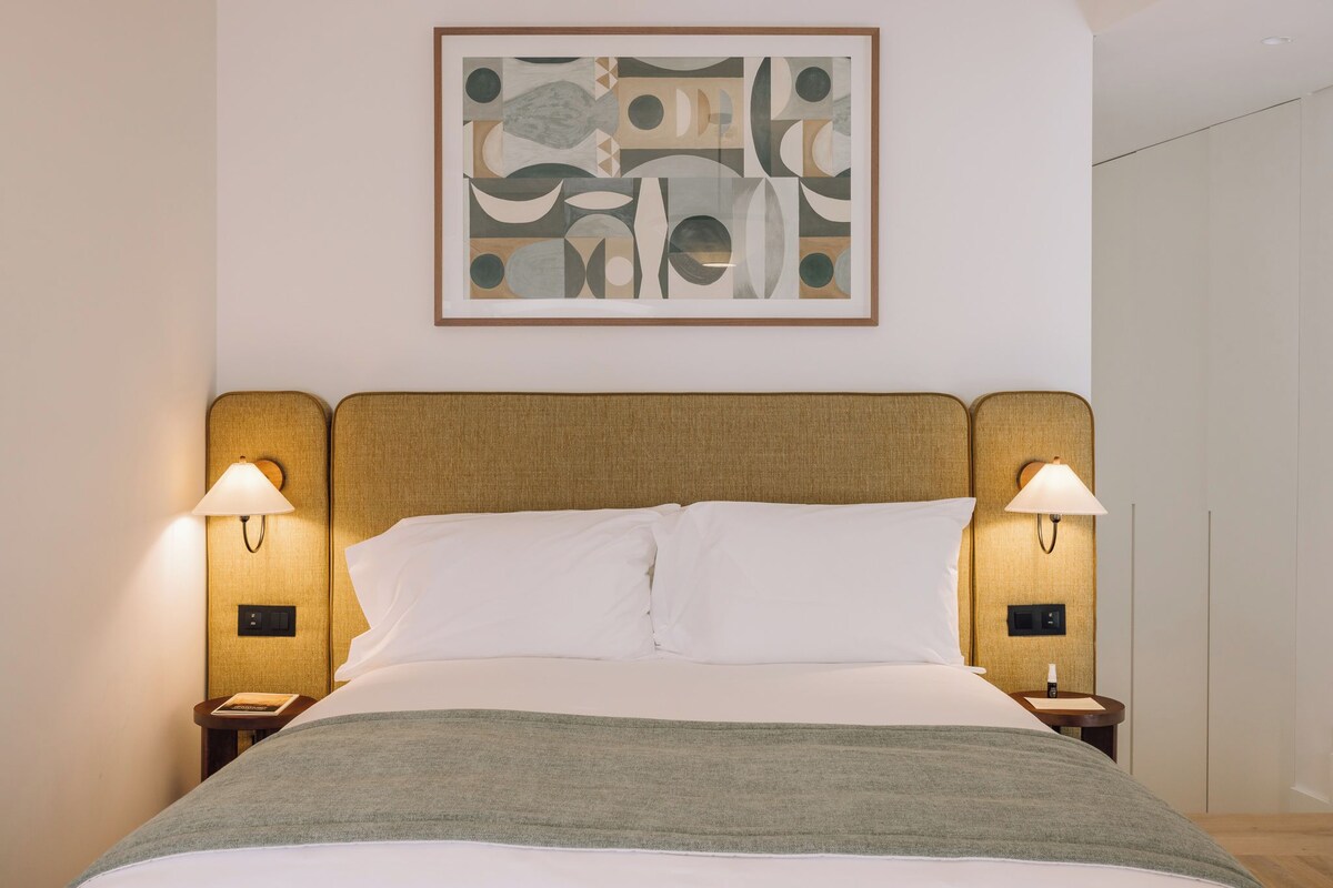 A neatly made bed with white linens and a grey throw, flanked by two bedside tables with lamps, in a room with a simple, modern design.
