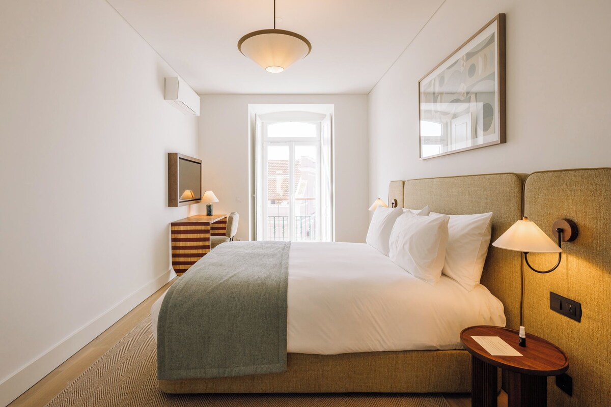 A hotel room with a bed, bedside tables, a desk with a chair, a large window, and a hanging light fixture.